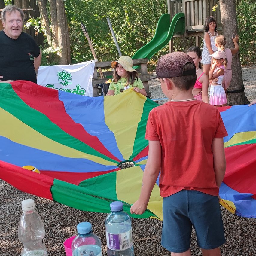 Eine Gruppe von Kindern und Erwachsenen spielt mit einem bunten Fallschirm in einem Park. Ein Junge in einem roten T-Shirt hält den Rand des Fallschirms.