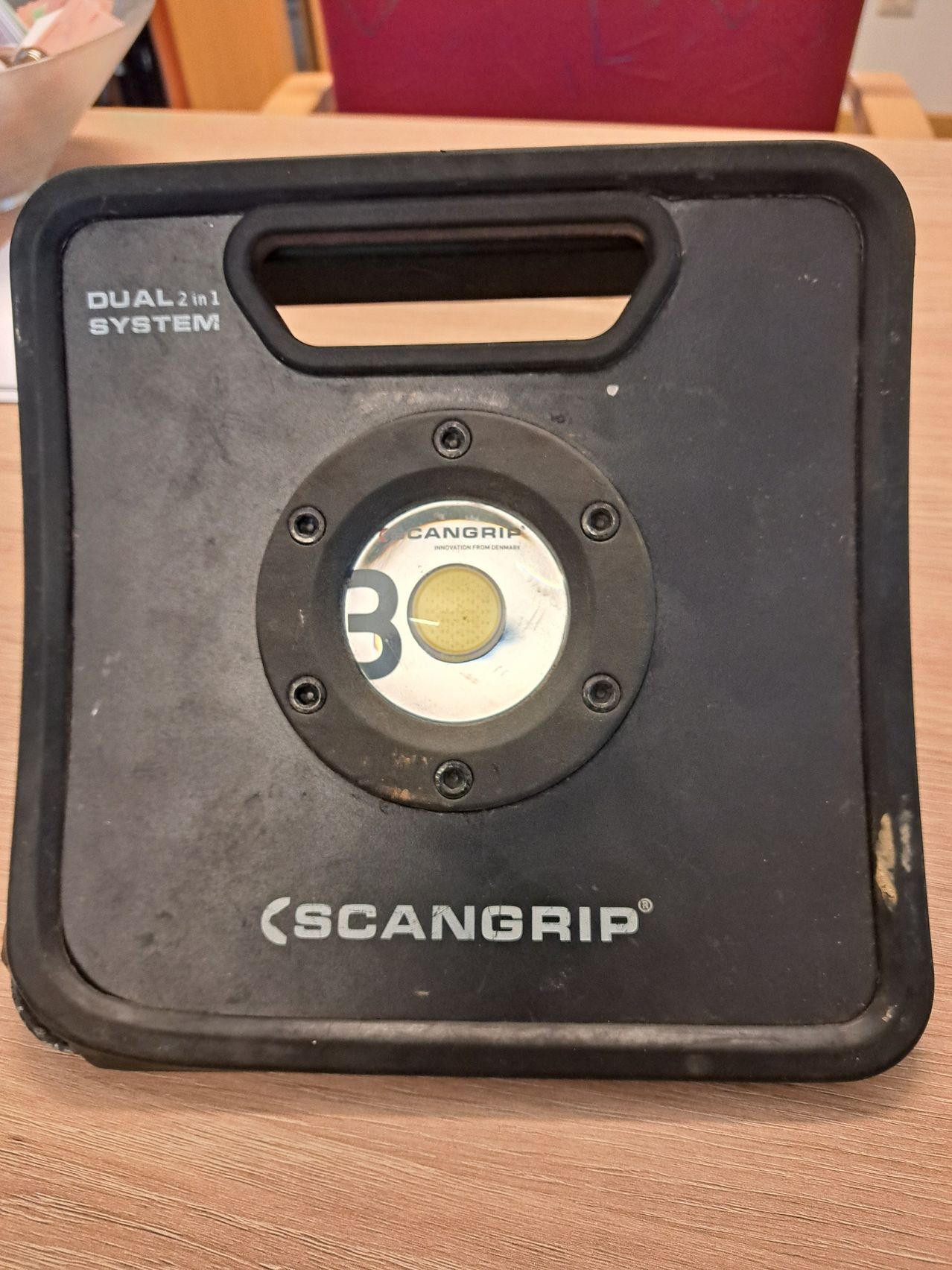 A black Scangrip dual 2 in 1 system with a yellow light on the front. The device is placed on a wooden surface.
