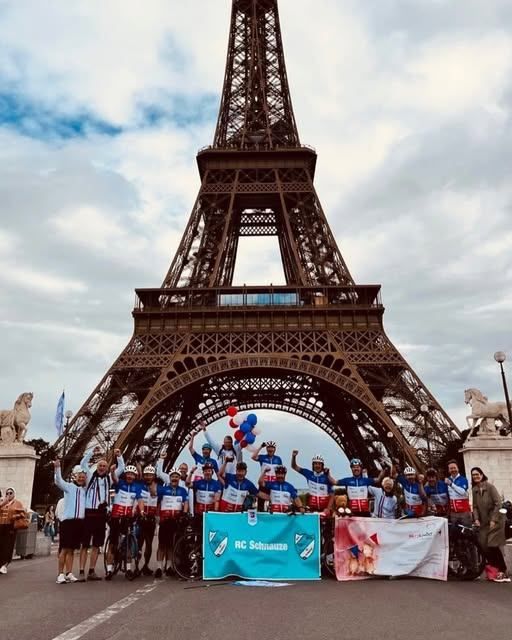 A group of cyclists pose for a photo in front of the Eiffel Tower, with a blue banner and balloons in the foreground.
