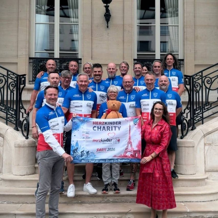 A group of people in cycling jerseys pose with a banner in front of a building. A woman in a red dress holds a teddy bear. The banner reads 'Herzkinder Charity Paris 2026'.