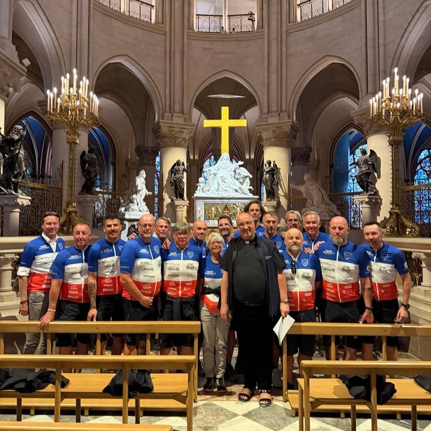 A group of cyclists wearing matching blue and red jerseys poses in a church with a priest and wooden pews.