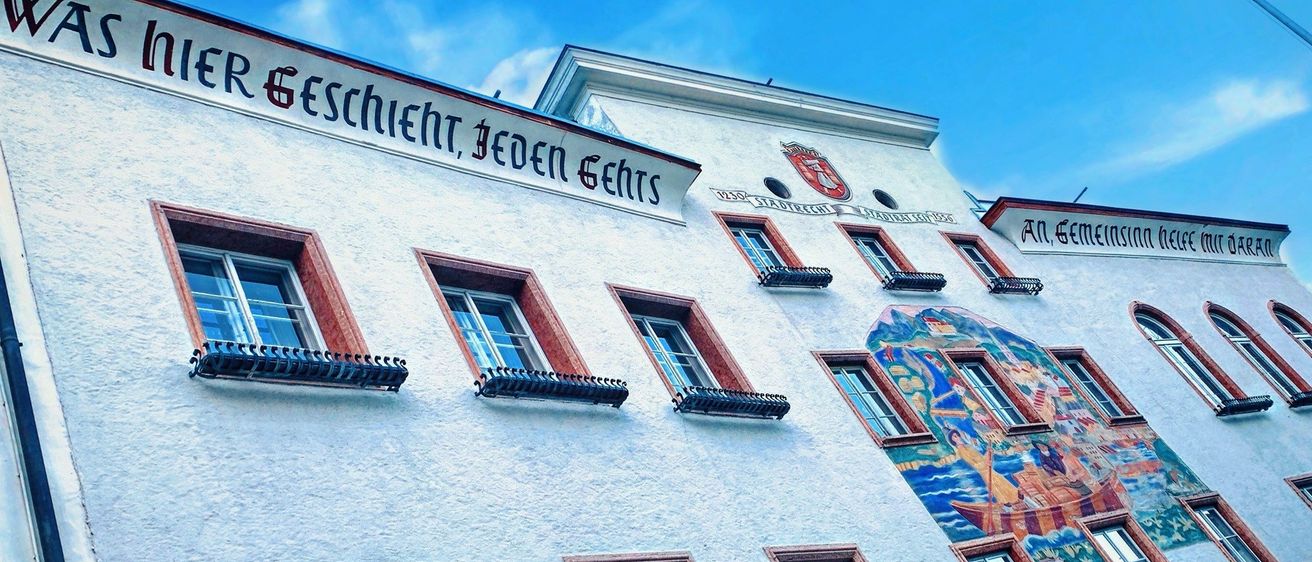 Ein weißes Gebäude mit rotem Rand und mehreren Fenstern zeigt einen deutschen Satz in großen Buchstaben. Der Text lautet 'Eschient, jeden gehrs'. Das Gebäude hat auch eine Wandmalerei eines Bootes an der Seite.