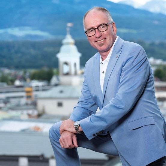 Ein lächelnder Mann in einem blauen Anzug und mit Brille lehnt sich gegen ein Geländer, mit einer Stadtsilhouette im Hintergrund.