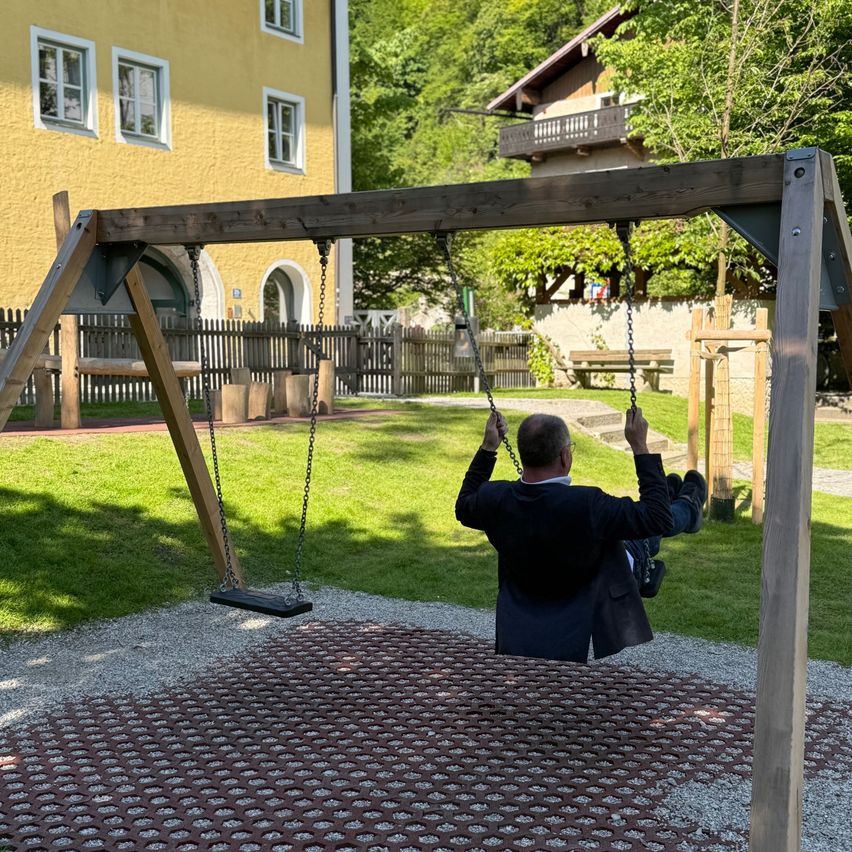 Ein Mann in einem Anzug schwingt auf einer Holzschaukel in einem Park mit einem gelben Gebäude im Hintergrund.