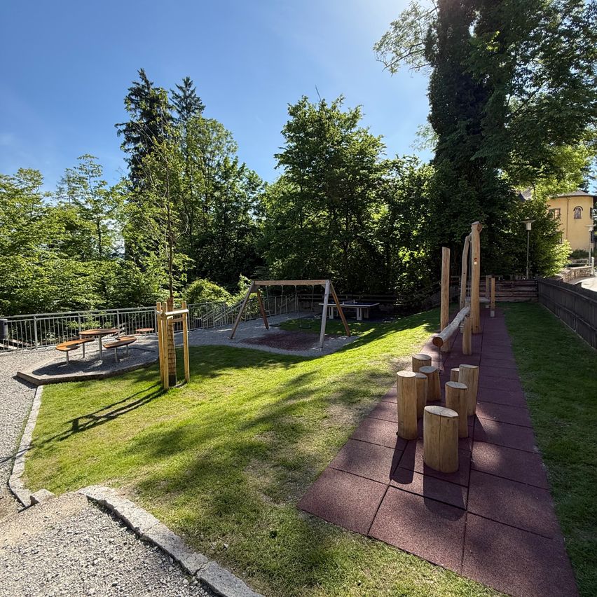 Ein Spielplatz mit Schaukel, Wippe und Klettergerüst. Der Bereich ist von Bäumen umgeben und hat einen Kiesweg. Unter dem Schatten steht ein Picknicktisch.