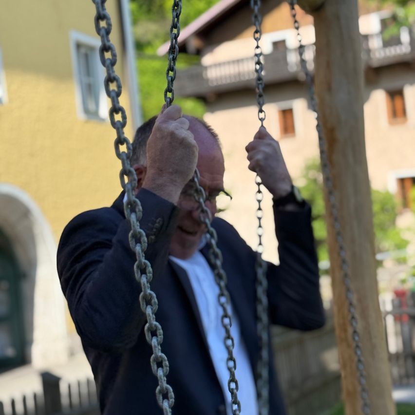 Ein Mann in Anzug und Brille lächelt, während er sich an einer Schaukelkette festhält. Dahinter befindet sich ein gelbes Gebäude mit einem bogenförmigen Eingang.