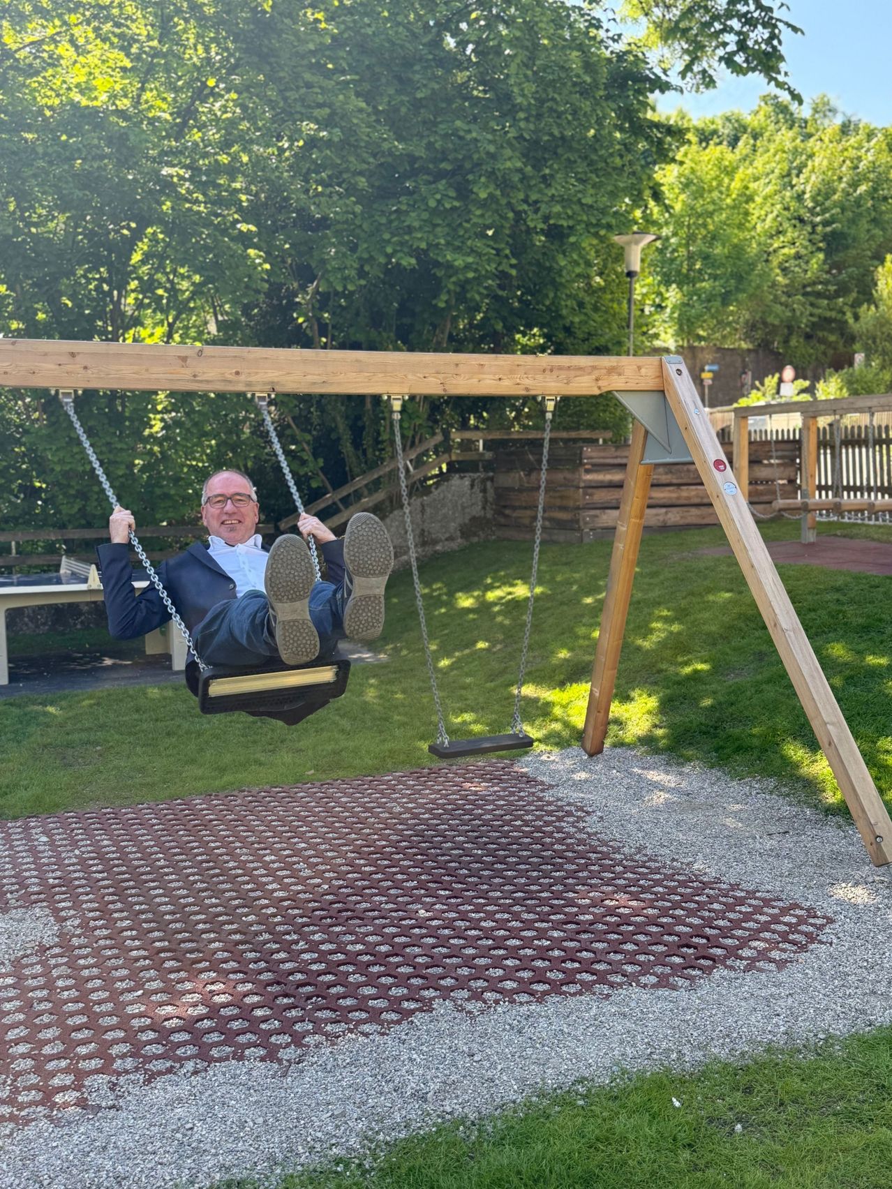 Ein Mann mit Brille und Anzug schwingt auf einer Holzschaukel in einem Park. Darunter befindet sich Kies und Gras. Hinter ihm steht ein Holzzaun.