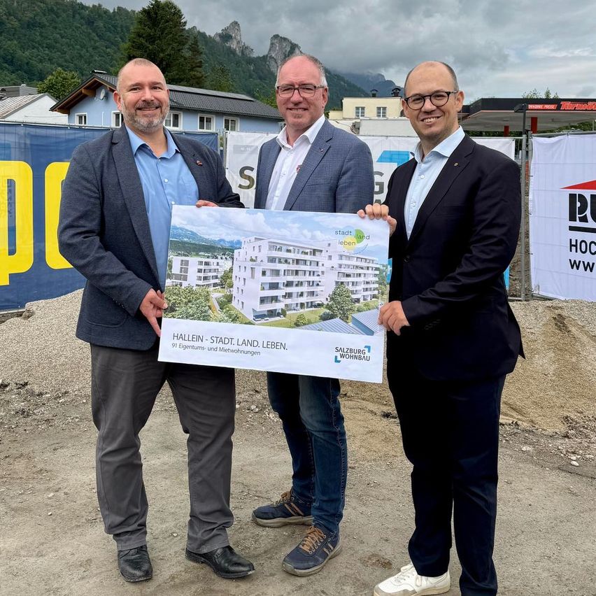 Drei Männer stehen lächelnd auf einer Baustelle. Sie halten ein Schild mit einem architektonischen Entwurf, auf dem 'Hallein - Stadt. Land. Leben.' steht. Hinter ihnen sind Banner mit Logos und Text zu sehen, und im Hintergrund sind Berge und Gebäude erkennbar.