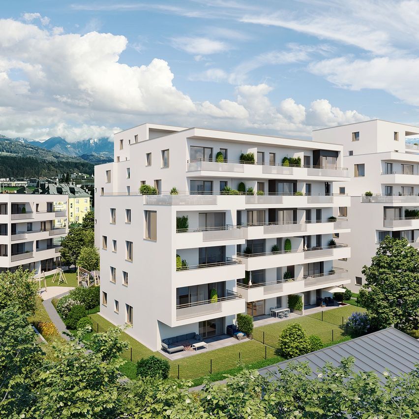 Luftaufnahme eines modernen weißen Wohngebäudes mit mehreren Balkonen und Grünflächen, in einer ruhigen Landschaft mit Bergen und blauem Himmel.