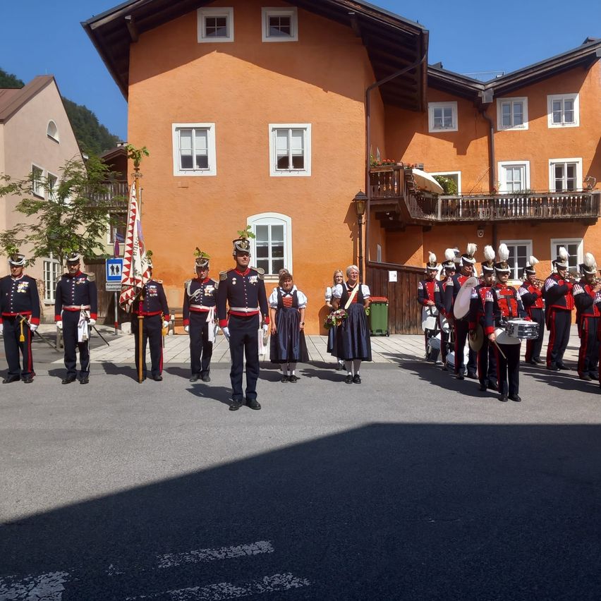 Eine Militärkapelle steht vor einem orangefarbenen Gebäude, mit einem Fahnenmast und einem Mülleimer in der Nähe. Die Musiker tragen Uniformen und Hüte, und einige halten Instrumente. Personen in traditioneller Kleidung sind in der Nähe.