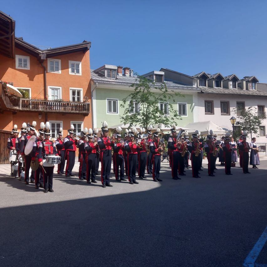 Eine Blaskapelle steht in einer Reihe auf einem Stadtplatz, gekleidet in rote und schwarze Uniformen und hält Musikinstrumente. Im Hintergrund befinden sich Gebäude mit Balkonen und Bäume.