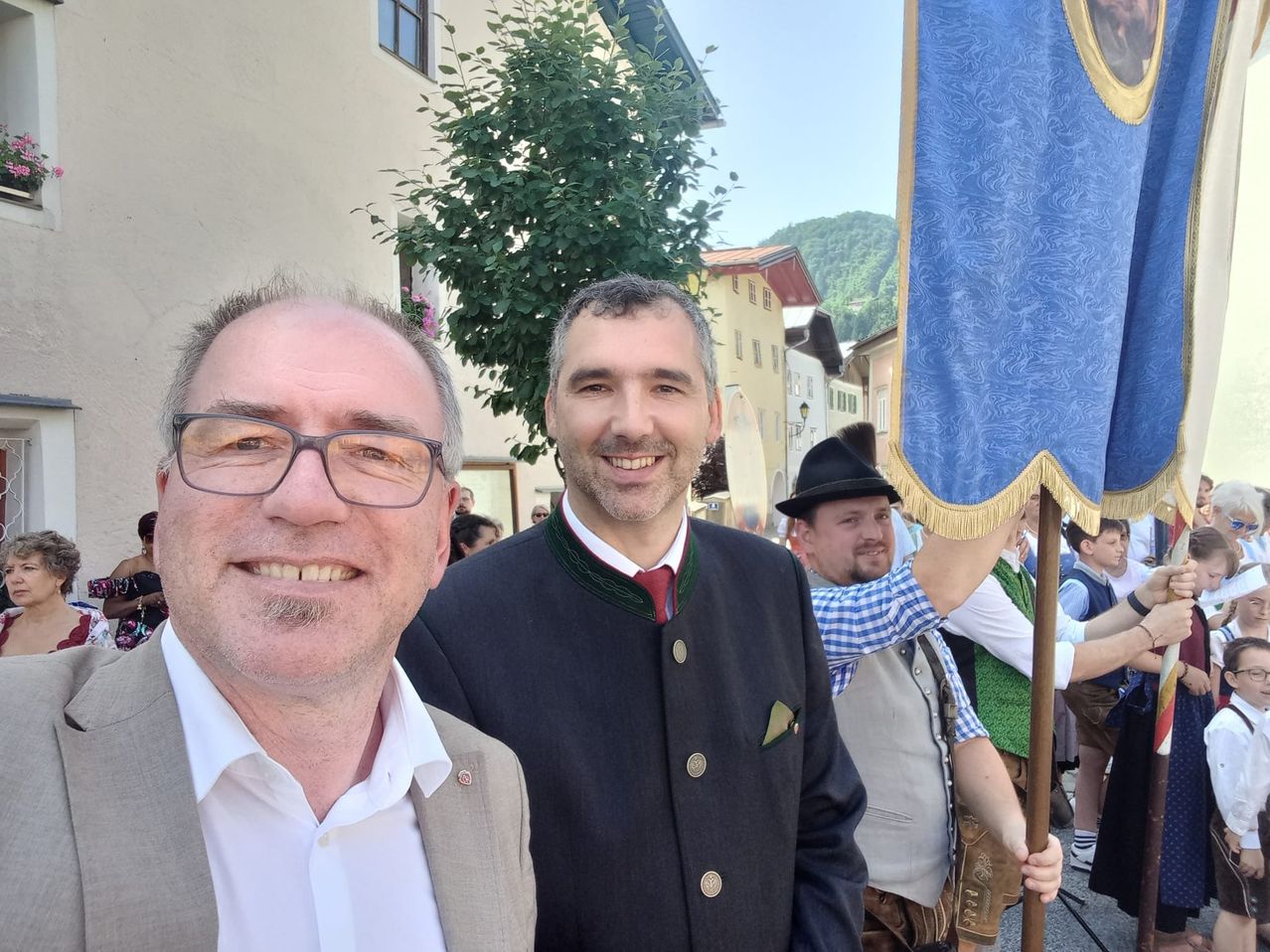 Zwei Männer lächeln für ein Foto, einer in formeller Kleidung, der andere in traditioneller deutscher Tracht. Hinter ihnen hält ein Mann eine blaue Fahne. Gebäude und ein Baum sind im Hintergrund zu sehen.