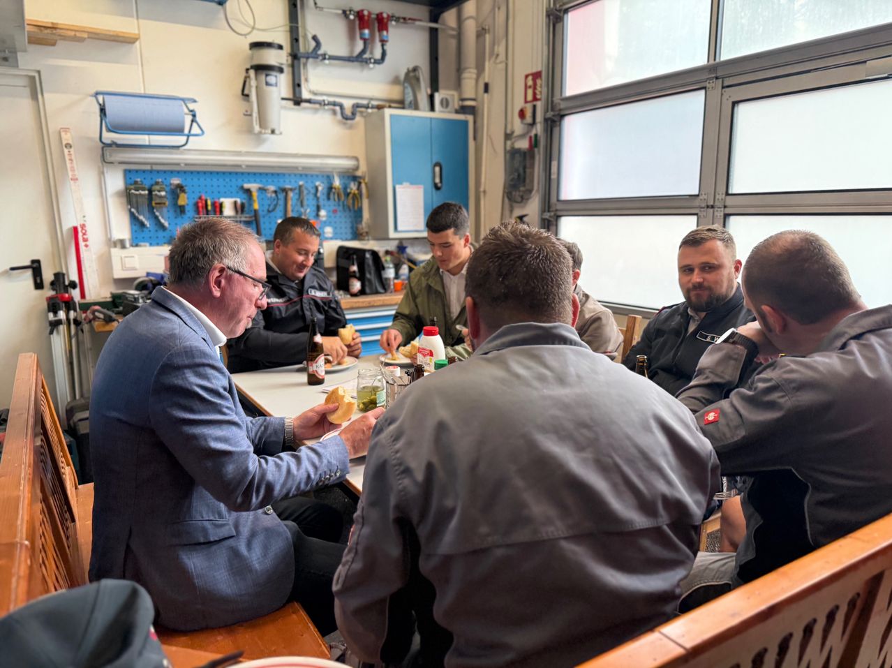 Mehrere Männer sitzen um einen Tisch in einer Werkstatt und genießen eine Mahlzeit. Ein Mann trägt einen Anzug, während andere lässige Kleidung tragen. Sie haben Flaschen, Gläser und Essen auf dem Tisch. Der Raum hat Regale mit Werkzeugen und ein großes Fenster.