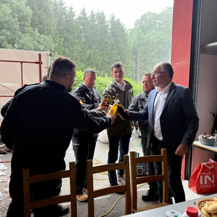 Mehrere Männer stehen draußen in der Nähe einer Tür. Ein Mann in einem Anzug gibt einem anderen Mann in Uniform eine Flasche. Bäume und ein weißer Lkw sind im Hintergrund.