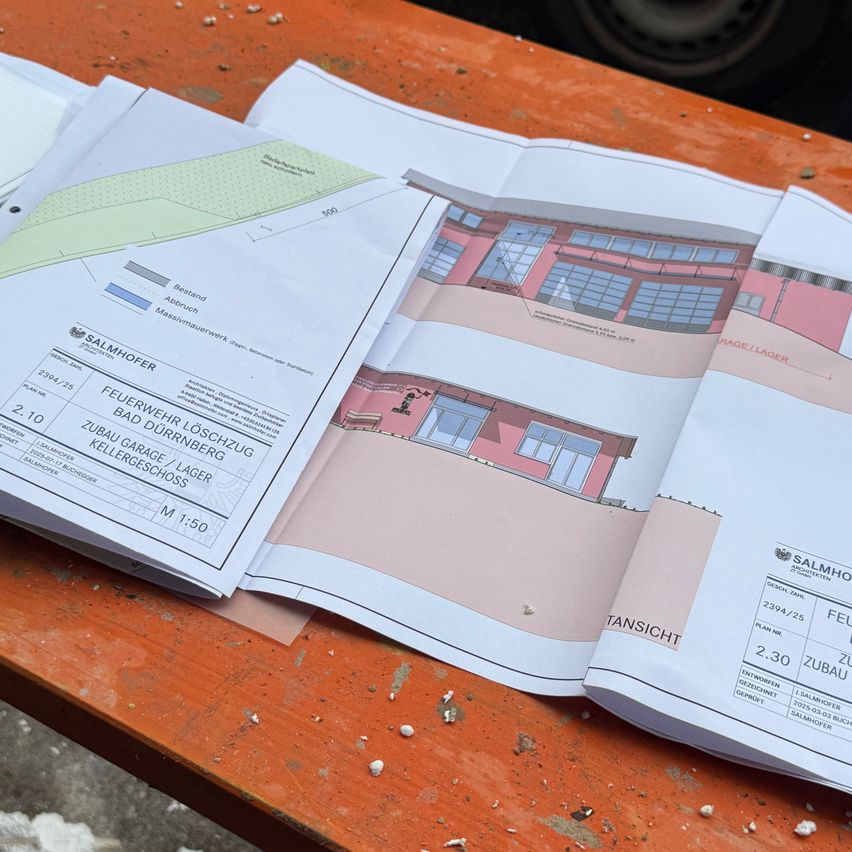 Ein Bauplan für ein Gebäude liegt auf einem Tisch. Er enthält eine Zeichnung eines Hauses mit mehreren Räumen, einschließlich Keller und Garage. Der Tisch besteht aus Holz.