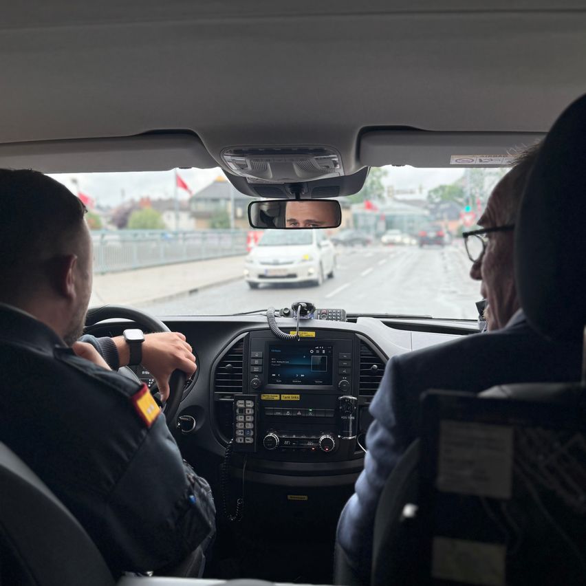 Zwei Männer in Polizeiuniformen sitzen im Rücksitz eines Fahrzeugs mit freier Sicht auf die Straße vor ihnen und einem weißen Auto im Rückspiegel.