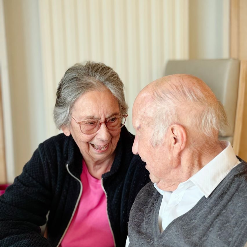 Eine ältere Frau mit Brille und rosa T-Shirt sitzt neben einem älteren Mann in weißem Hemd und grauem Pullover, beide lächeln und schauen sich an.