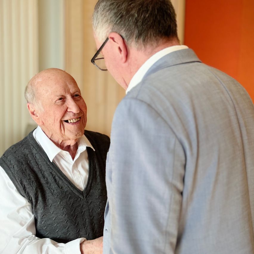 Ein älterer Mann mit grauer Weste lächelt, während er mit einem Mann in einer grauen Anzugjacke und einer Brille spricht.