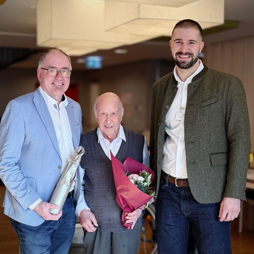 Drei Männer stehen lächelnd zusammen. Einer hält einen Blumenstrauß, der andere ein eingewickeltes Geschenk. Sie sind in einem Raum mit Holzboden und einer Treppe.