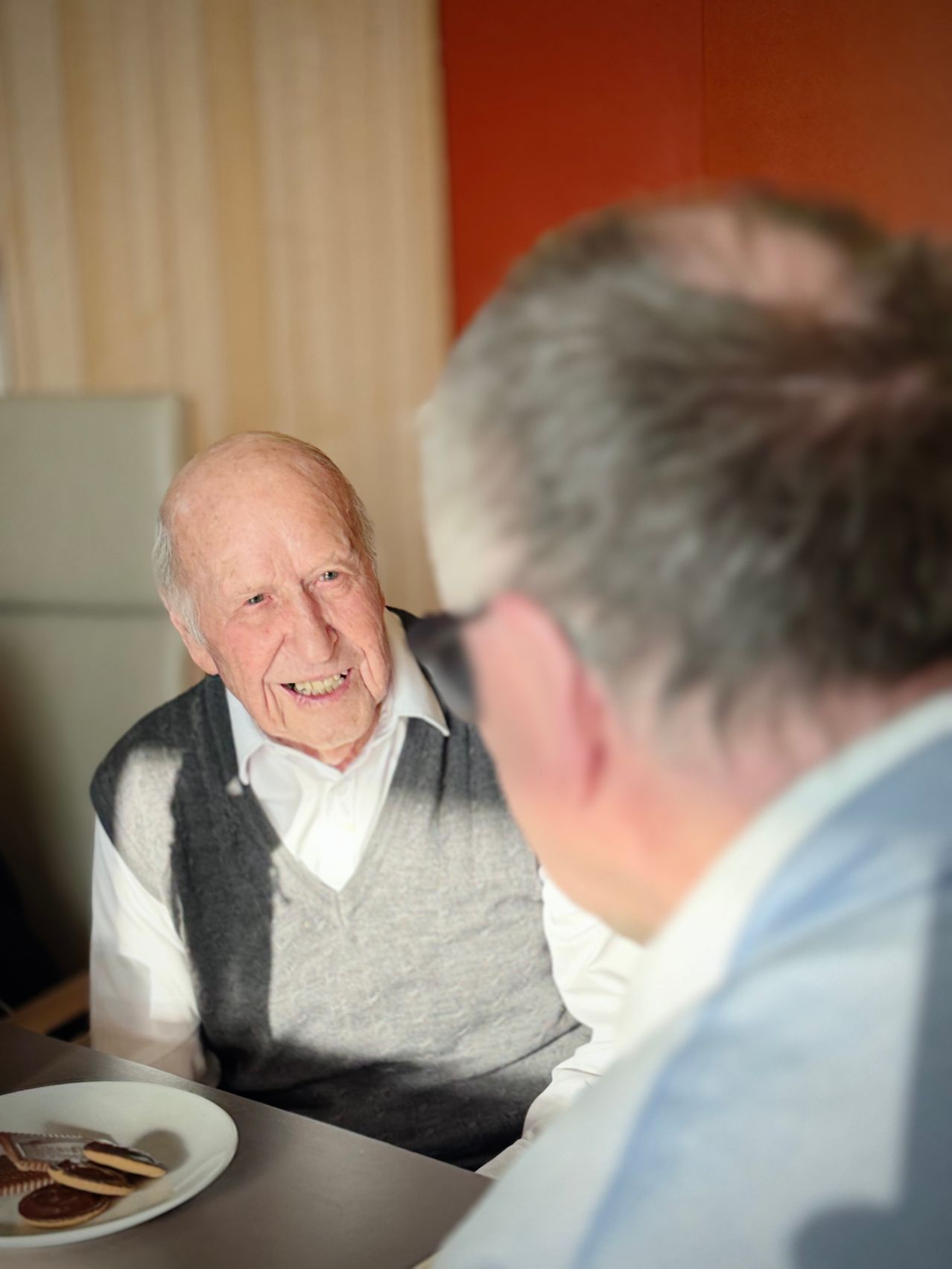 Ein älterer Mann mit grauem Weste und Lächeln sitzt und unterhält sich mit einem anderen Mann mit Brille. Der Ort scheint ein Zimmer mit Holzwänden zu sein.