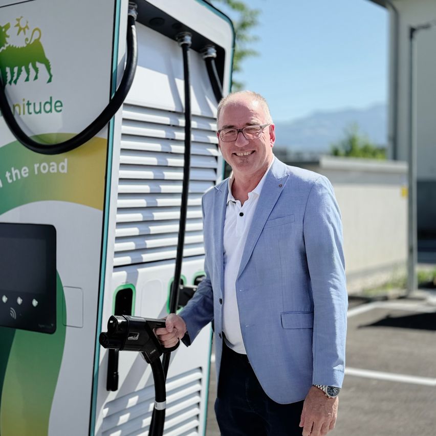 Ein Mann in einem blauen Anzug steht neben einer Ladestation für Elektrofahrzeuge, lächelt und hält den Ladekabel.