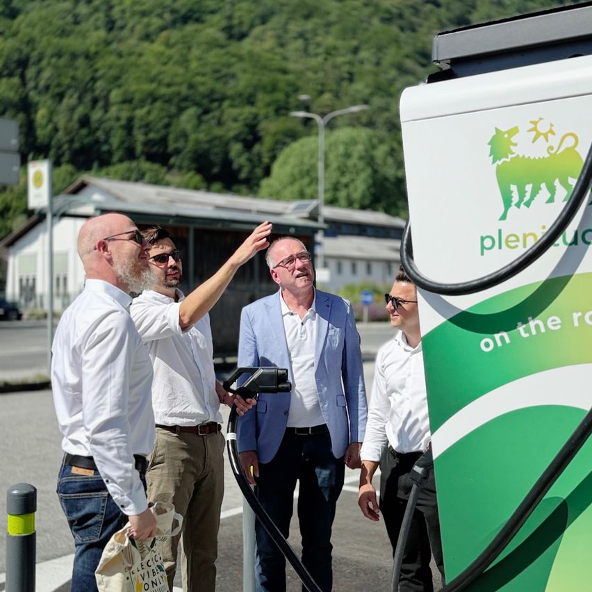 Vier Männer stehen in der Nähe einer Plenitude-Ladestation für Elektrofahrzeuge. Ein Mann hält einen Ladekabel, während ein anderer auf die Station zeigt.
