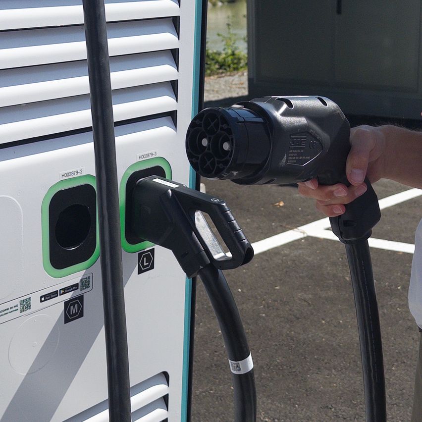 Eine Person steckt einen schwarzen Ladekabel in eine Ladestation für Elektrofahrzeuge. Die Station verfügt über mehrere Ladepunkte, von denen einer gerade in Gebrauch ist.