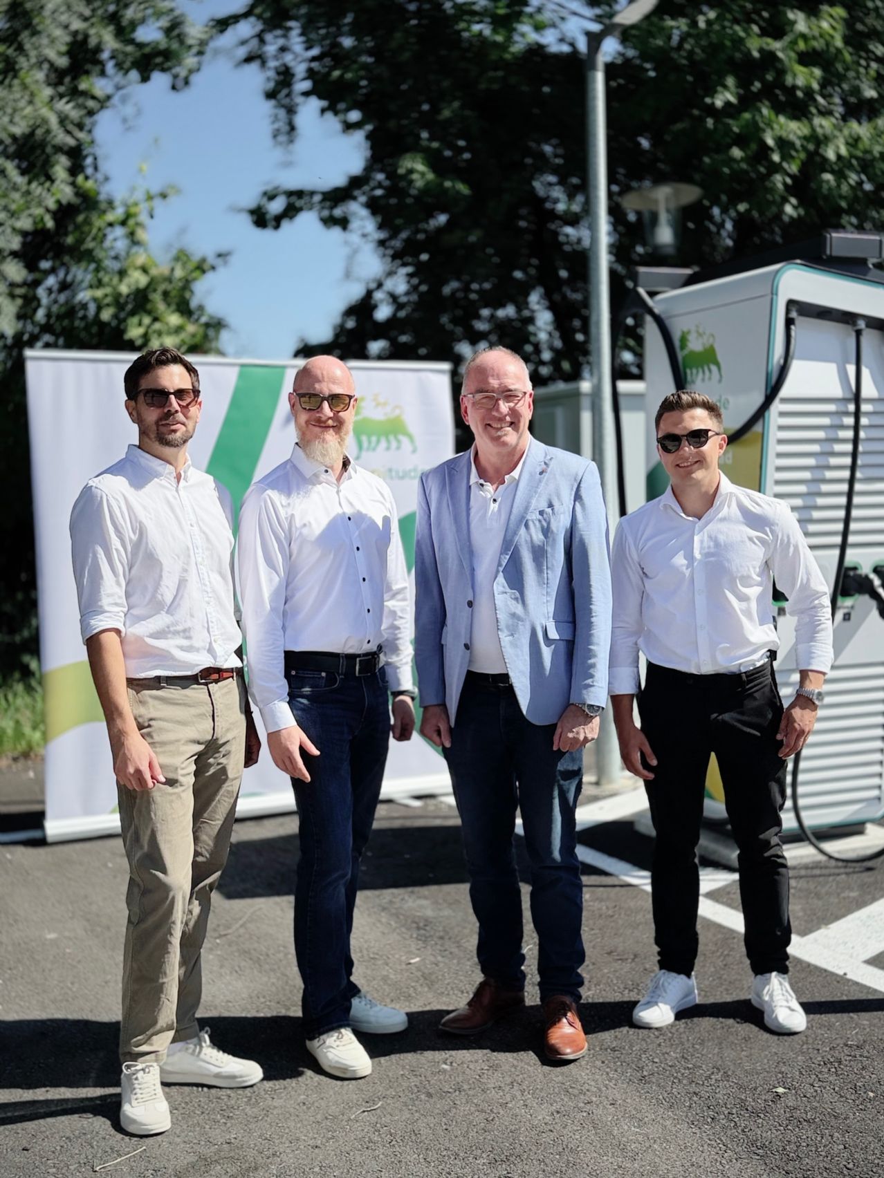 Vier Männer in formeller Kleidung stehen vor einer Ladestation für Elektrofahrzeuge, einer der Männer trägt einen hellblauen Blazer.
