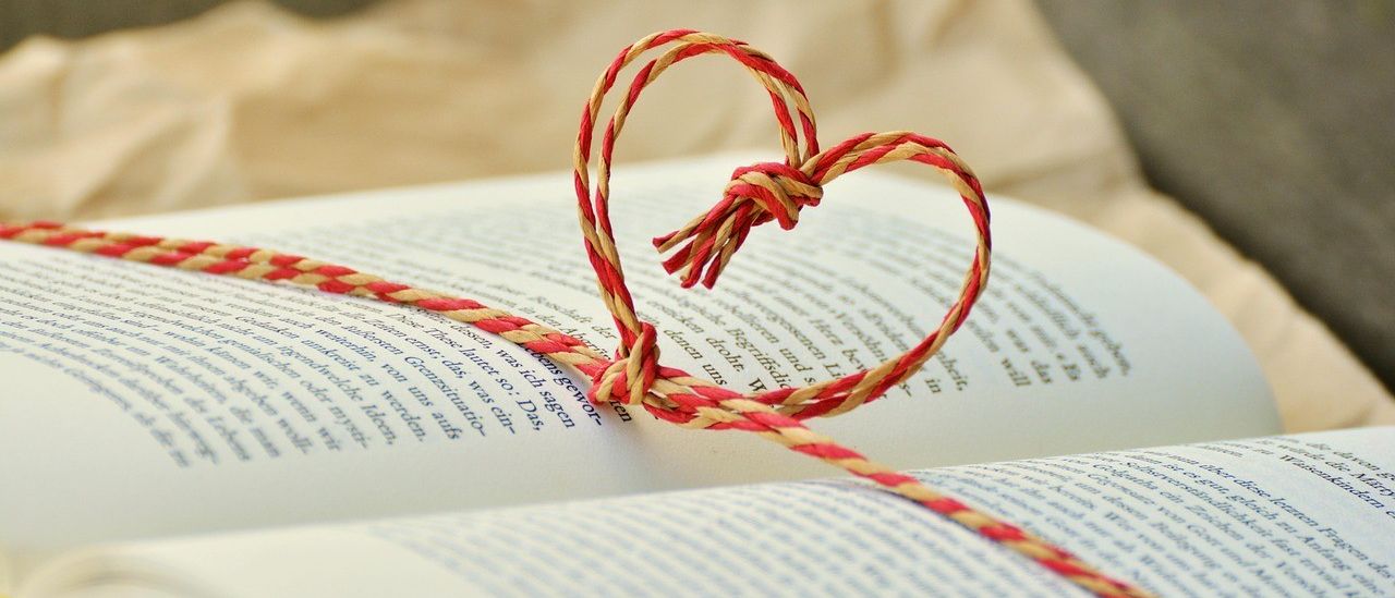 Ein herzförmiger Band-Lesezeichen ist in einem offenen Buch gebunden. Das Buch ist leicht geöffnet, mit unschärfem Text im Hintergrund.