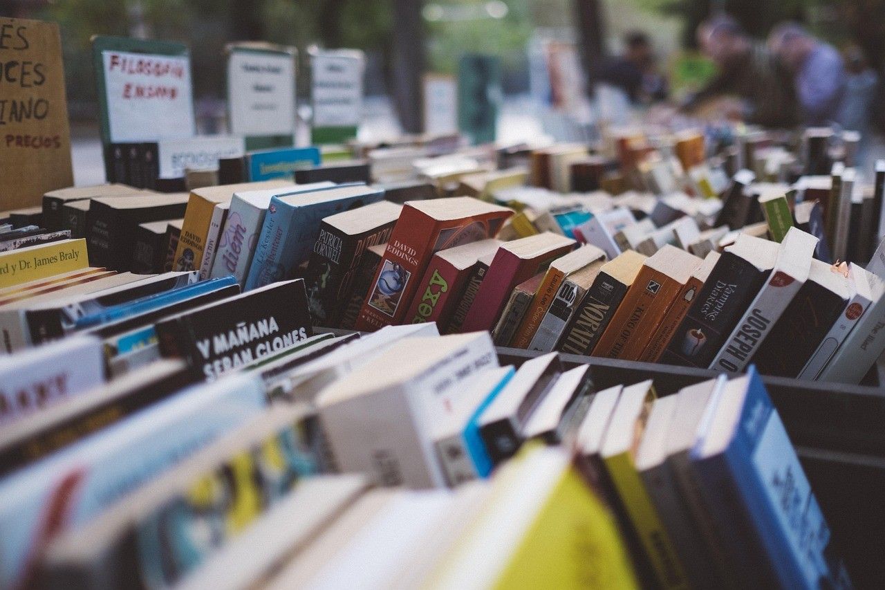 Ein großer Haufen Bücher wird in einer Außenumgebung ausgestellt. Verschiedene Bücher in unterschiedlichen Größen und Farben sind aufeinandergestapelt. Im Hintergrund sind einige Personen zu sehen.
