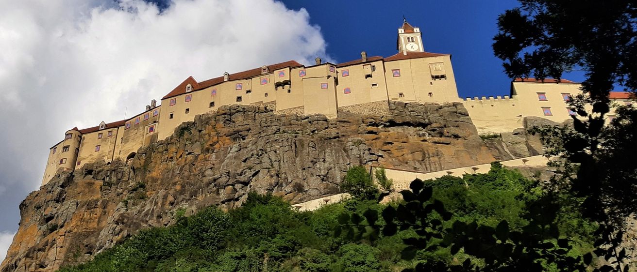 Eine Burg auf einem Felsvorsprung mit einem bewölkten Himmel darüber. Die Burg hat einen Glockenturm und ist von Grün umgeben.