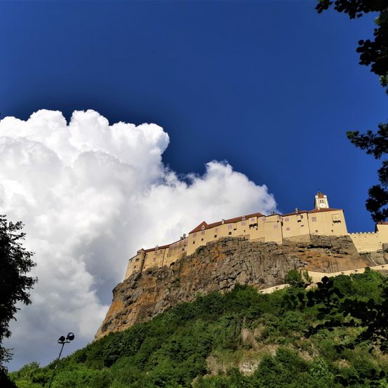 Eine Burg auf einem Felsen mit einer großen Wolke am Himmel und Bäumen darunter.