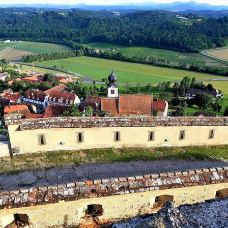 Luftaufnahme eines historischen Dorfes mit einer Kirche, umgeben von üppigen grünen Feldern und Bergen in der Ferne.
