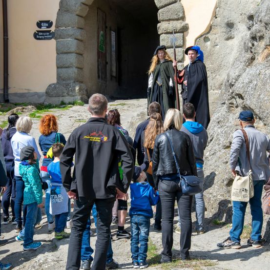 Eine Gruppe von Menschen steht vor einem historischen Gebäude und beobachtet zwei Personen in Kostümen. Einer hält eine Lanze, während der andere einen Hut trägt.