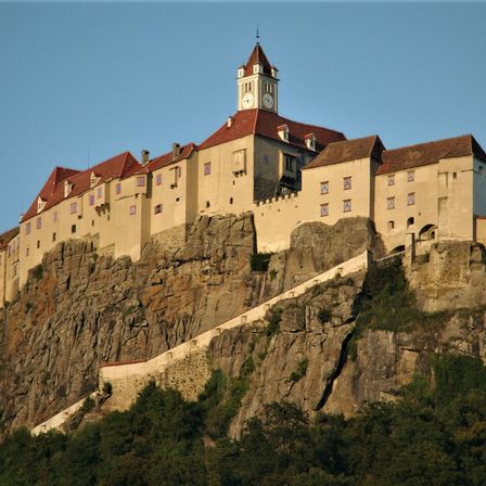 Eine Burg mit Uhrenturm steht auf einem steilen Felsvorsprung unter klarem Himmel, umgeben von Grün.