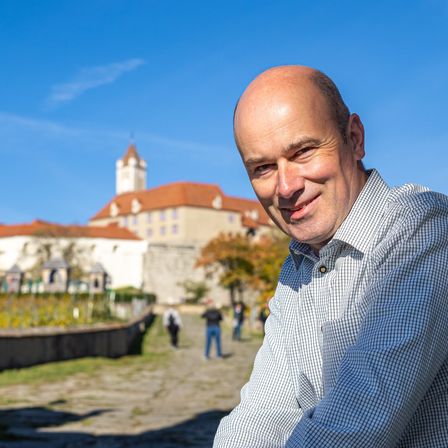 Ein lächelnder Mann steht vor einem historischen Gebäude unter blauem Himmel. Im Hintergrund gehen Menschen vorbei.