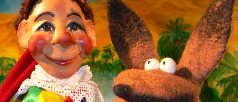 Eine Puppe mit rotem Hut steht neben einem Holzkasten voller Gold. Die Puppe trägt ein farbenfrohes Kostüm.