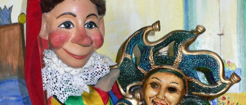 Eine Puppe mit rotem Hut hält eine bunte Maske mit lächelndem Gesicht, vor einem gemalten Hintergrund.