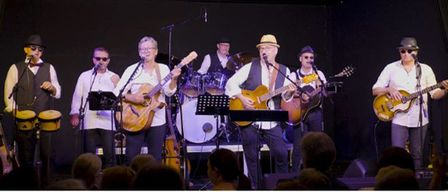 Eine Band spielt auf der Bühne mit Gitarren, einem Schlagzeug und einer Tambourin. Mehrere Zuschauer sind im Vordergrund sichtbar.