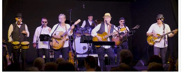 Eine Band spielt auf der Bühne mit Gitarren, einem Schlagzeug und einer Tambourin. Mehrere Zuschauer sind im Vordergrund sichtbar.