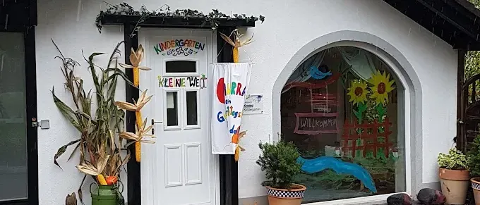 Die Vorderseite eines Kindergartens zeigt eine weiße Tür mit bunten Buchstaben und einem kleinen Garten mit Pflanzen, Kürbissen und Mais. Ein Banner hängt über der Tür, und ein Glasfenster zeigt eine Zeichnung eines Vogels und Sonnenblumen.