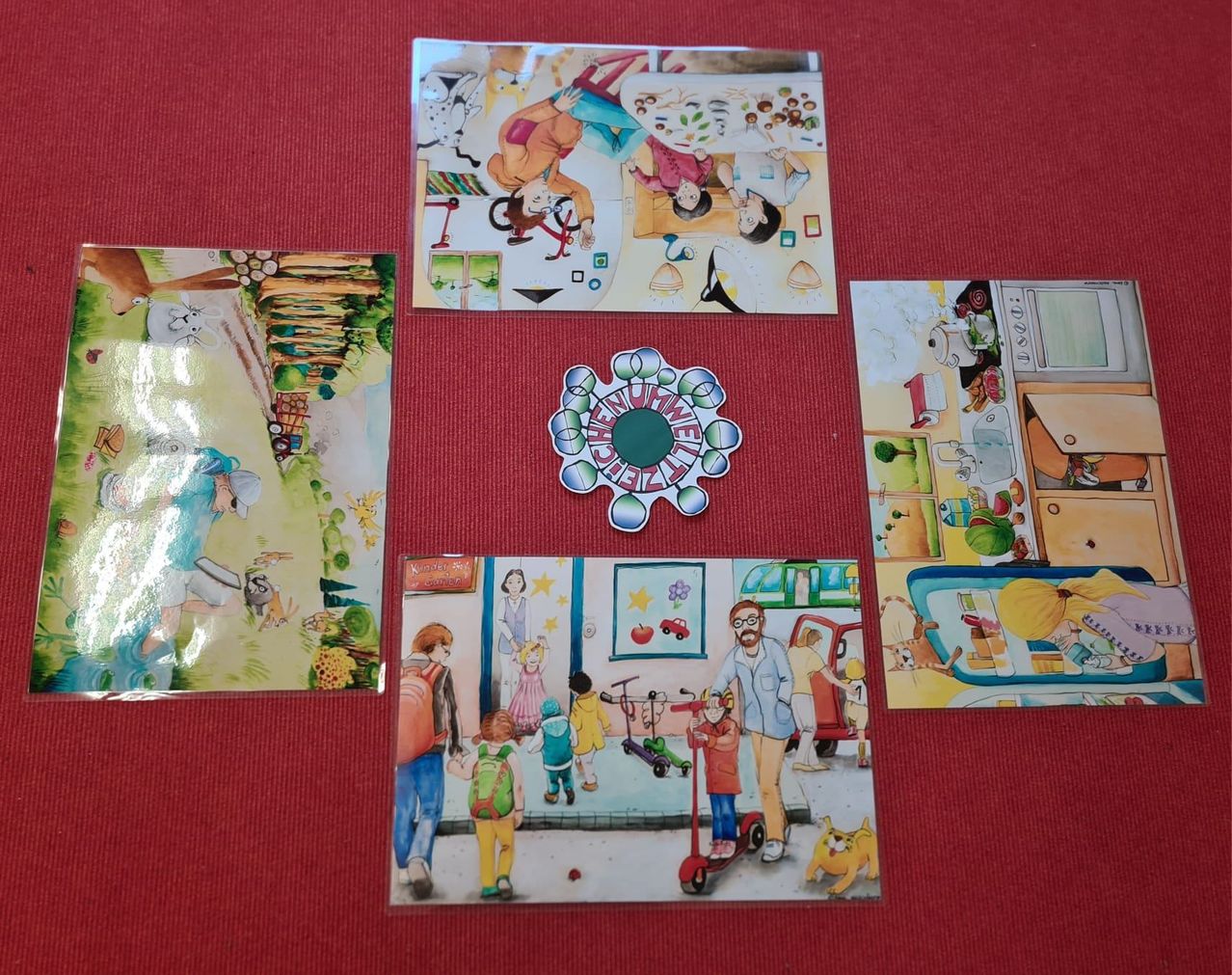 A collection of colorful children's book pages and a sticker are displayed on a red surface. The pages depict various scenes, including a person riding a bicycle, a family in a kitchen, and children walking with a man. The sticker has a circular design with text.