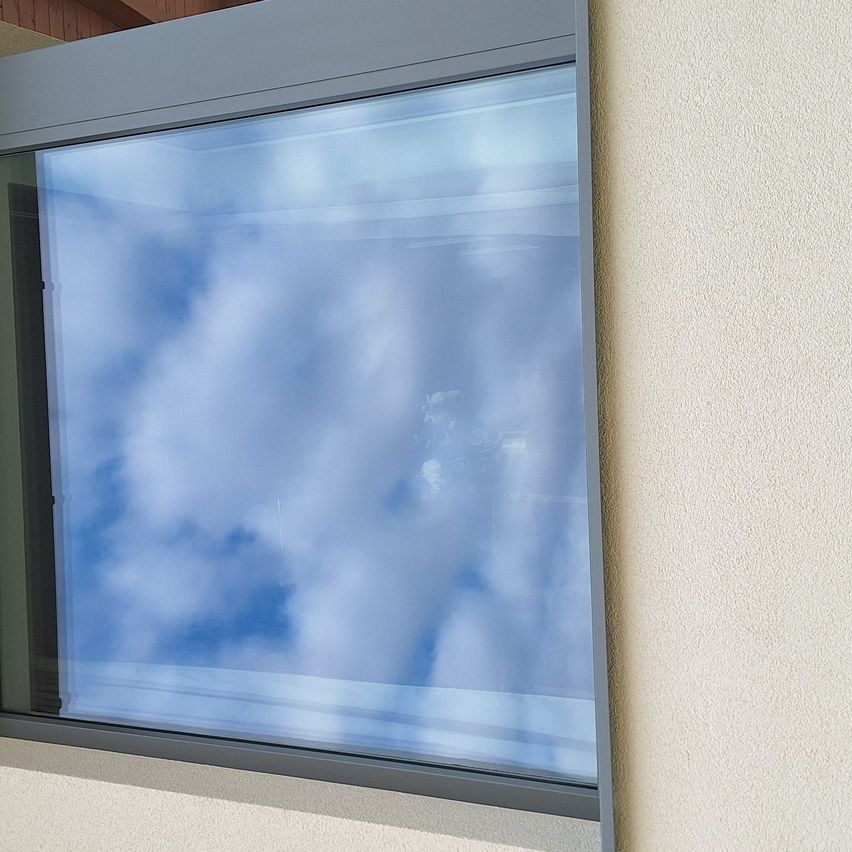 Ein Fenster mit grauem Rahmen ist an einer beigen Wand montiert und spiegelt einen blauen Himmel mit Wolken wider.