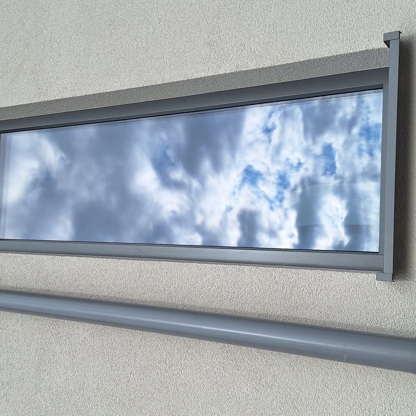 Ein Fenster mit grauem Rahmen ist an einer weißen Wand montiert und spiegelt den bewölkten Himmel wider.