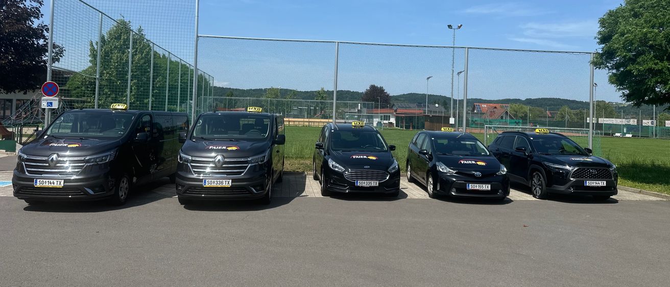 Fünf schwarze Autos, wahrscheinlich Taxis, sind in einer Reihe auf einem Parkplatz geparkt. Ein Metallzaun ist im Hintergrund zu sehen, und es sind Bäume und Berge in der Ferne sichtbar.