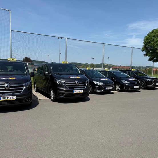 Mehrere schwarze Taxis sind auf einem Parkplatz hinter einem Zaun geparkt, mit einem klaren blauen Himmel im Hintergrund.