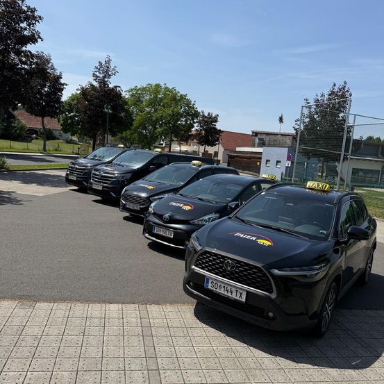 Fünf schwarze Autos sind auf einem Parkplatz geparkt, mit Bäumen und Gebäuden im Hintergrund. Die Autos haben gelbe Taxi-Zeichen obenauf.