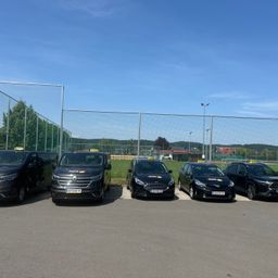 Fünf Taxis sind auf einem Parkplatz mit einem Zaun geparkt. Der Himmel ist blau mit einigen Wolken. Bäume und Straßenlaternen befinden sich hinter dem Zaun.