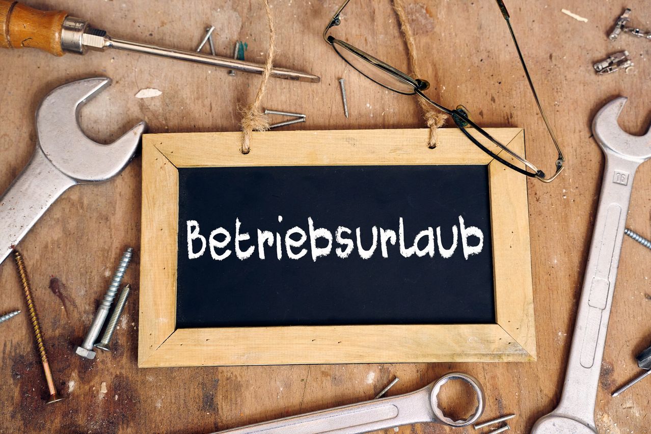 Ein Schultafel mit dem deutschen Wort Betriebsurlaub darauf, auf einer Holzoberfläche mit verschiedenen Werkzeugen und Gegenständen wie einem Hammer und Brille um sie herum.
