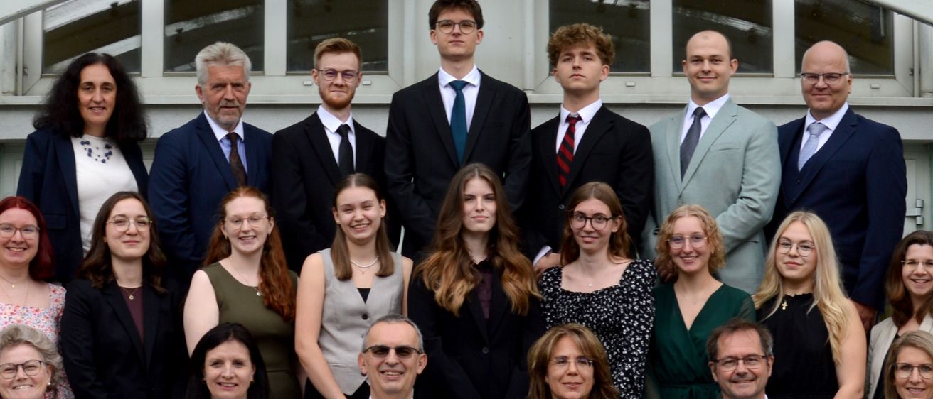 Eine Gruppe von Menschen, die feierlich gekleidet sind, sitzen und stehen in Reihen, wahrscheinlich für ein Foto. Einige lächeln. Sie befinden sich in einem Innenbereich mit einer weißen Wand und Glasfenstern.